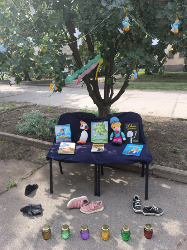 Під деревом стоїть лава. На ній лежать книги, іграшки. Біля лави - дитяче взуття, свічки.