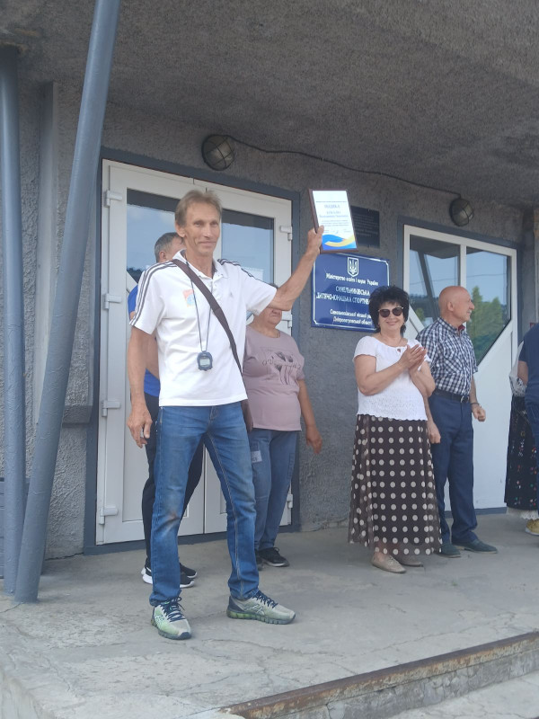 На ганку школи стоїть група чоловіків і жінка. Один із чоловіків - попереду інших. Тримає у руках грамоту. 