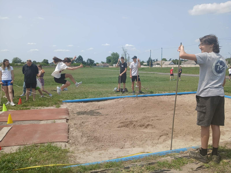 Яма для стрибків у довжину. На фото - момент стрибка дівчини. Поряд з ямою -  троє хлопців стоять із граблями, чоловік і жінка, інші діти.