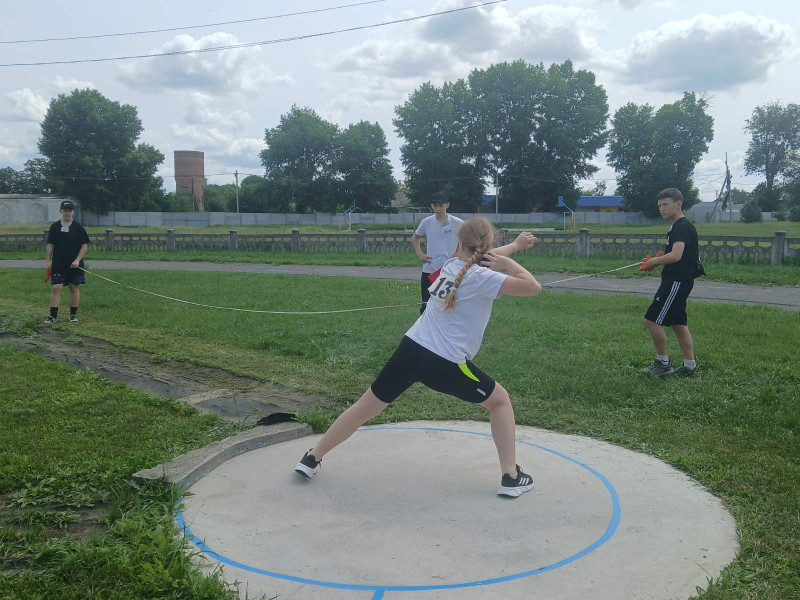 У колі стоїть юна спортсменка. Права рука - зігнута у лікті, тримає ядро. Ліва - виставлена  вперед. Неподалік - стоять  три юнака. Двоє натягнули вимірювальну стрічку.