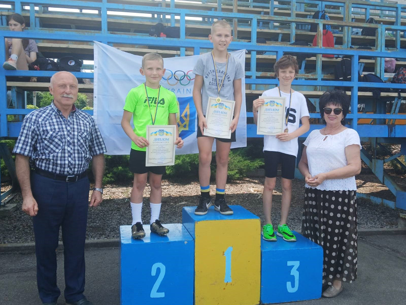 Троє юнаків стоять на п'єдесталі для нагородження з медалями і грамотами.  Праворуч стоїть жінка, ліворуч -чоловік.