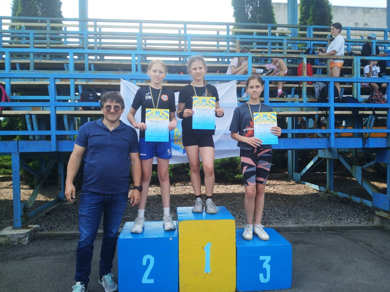 Троє дівчат стоять на п'єдесталі для нагородження з медалями і грамотами. Ліворуч  стоїть чоловік.