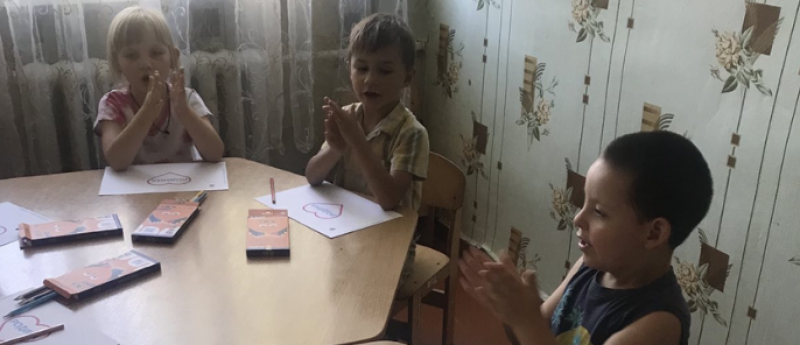 Дівчинка і два хлопчики сидять за столиком, на якому лежать олівці у коробках, аркуші паперу із сердечками.  Щось промовляючи, хлопають у долоньки.