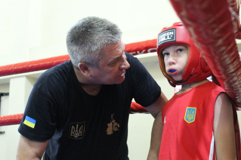 Тренер Олексій Решетняк, нахилившись, дає настанови юному боксеру, який стоїть  у куті рингу.