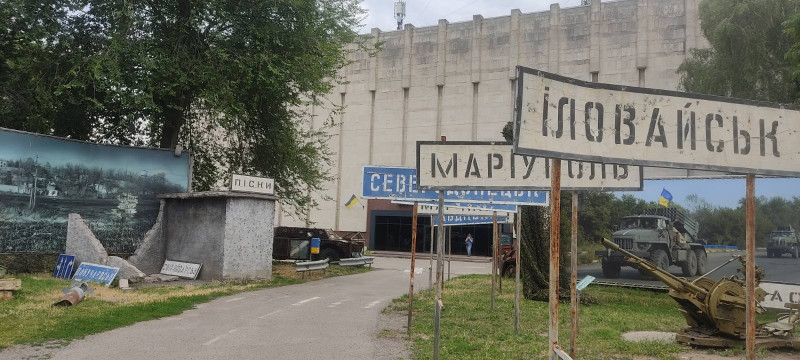 На задньому плані - будівля. До неї йде асфальтована дорога. Вздовж дороги праворуч стоять  дорожні знаки  з назвами населених пунктів: Іловайськ, Маріуполь, Северодонецьк та інші.  Ліворуч - залишки зупинки із табличкою "Піски". За нею -  велике фото у сірому тоні з краєвидом, уламки боєприпасів.  