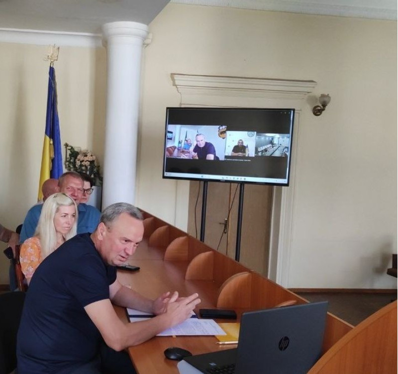 Міський голова Дмитро Зражевський разом із секретарем міської ради, заступниками, керуючою справами виконавчого комітету  сидять за столом перед екраном ноутбука. На задньому плані - великий екран із відеотрансляцією двосторонньої зустрічі.