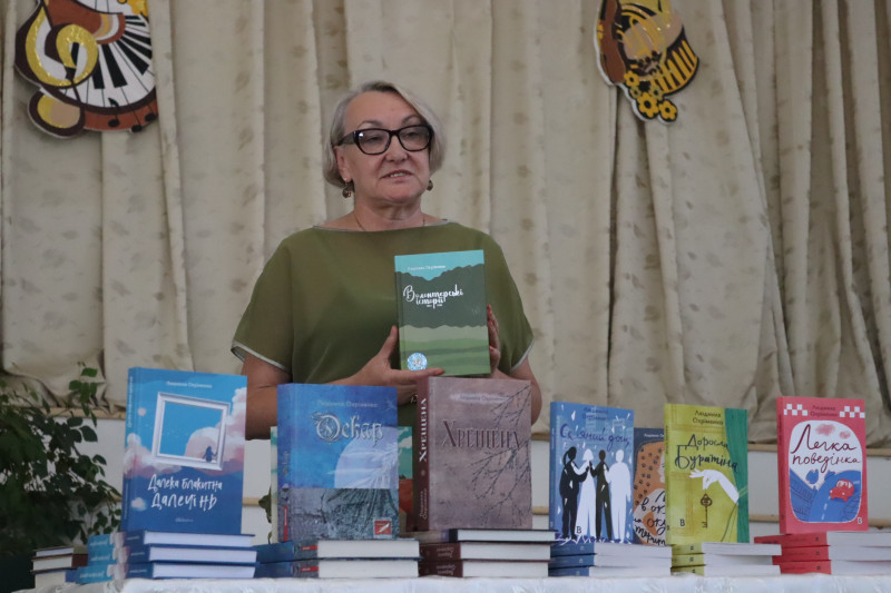 Письменниця Людмила Охріменко стоїть за столом з книгою  в руках. На столі - її авторські книги.