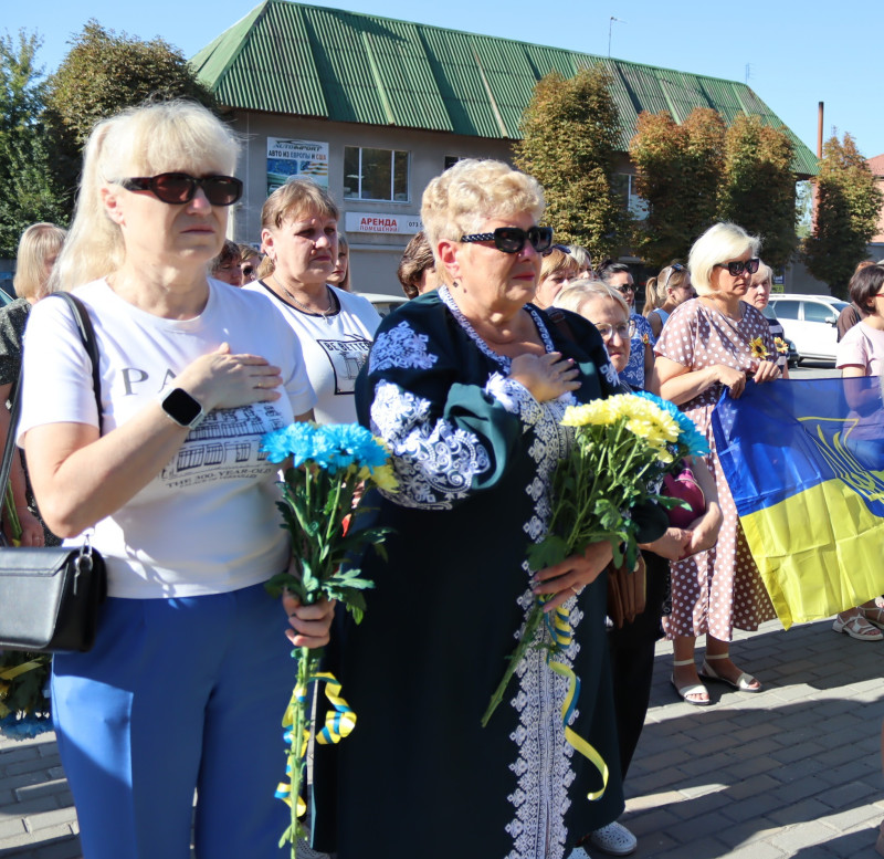 Жінки, різні за віком, стоять з букетами блакитно-жовтих квітів, інші тримають в руках  Прапор України. 