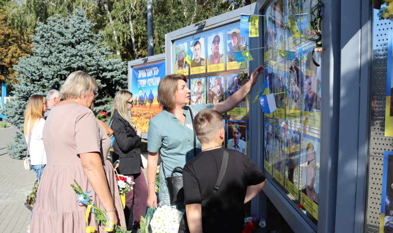 Жінки та хлопець стоять перед пам'ятною дошкою загиблим Героям. Мати показує дитині фото на стенді.