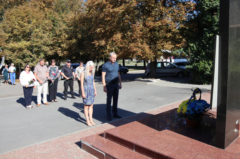 Міський голова Дмитро Зражевський разом із секретарем міської ради Оленою Заікою, іншими посадовцями, працівником поліції стоять перед пам'ятним знаком загиблим воїнам. У підніжжя пам'ятного знаку - кошик із квітами.