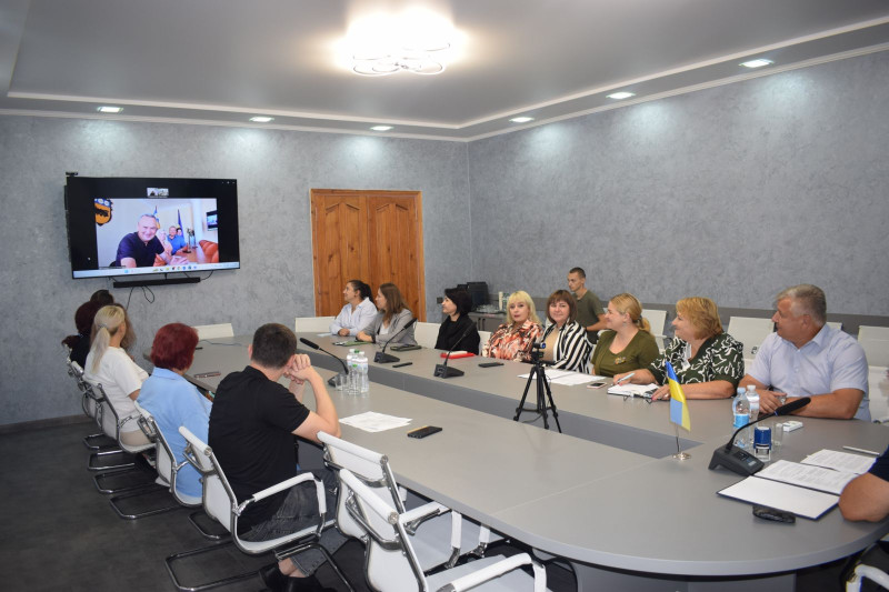 У кімнаті  за столом сидять чоловіки та жінки, погляди спрямовані на екран, розміщений на стіні.