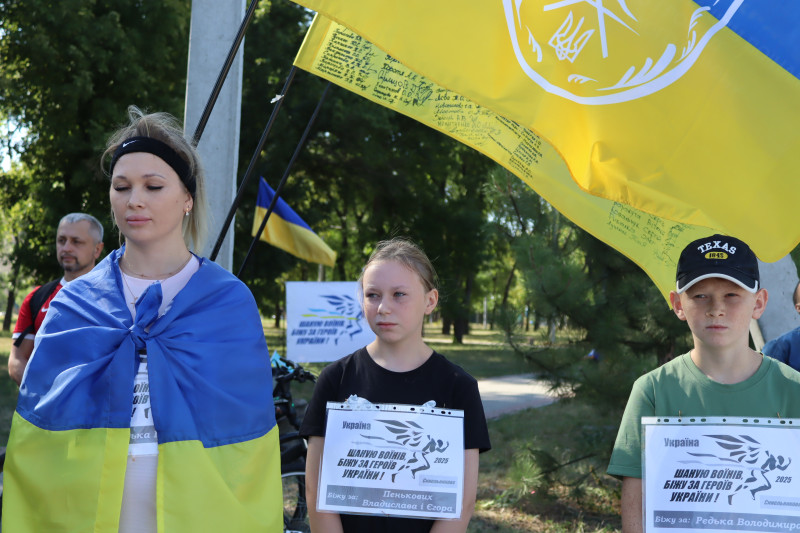 Молода дівчина, огорнута у Прапор України, дівчинка, хлопчик стоять на фоні прапорів, на футболках - прізвища Героїв.