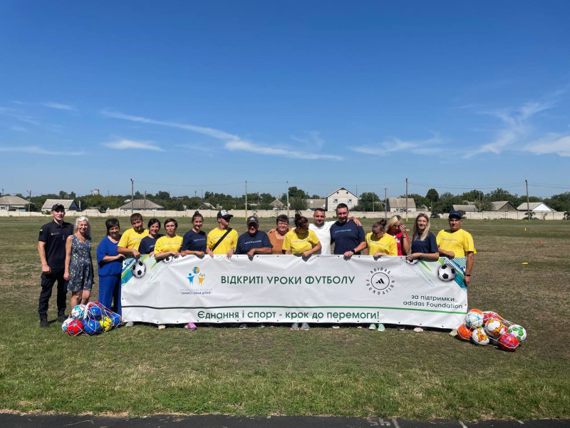 На зеленому полі спільне фото організаторів заходу, секретаря міської ради Олени Заіки, представника поліції  з банером: "Відкриті уроки футболу. Єднання і спорт - крок до перемоги". 