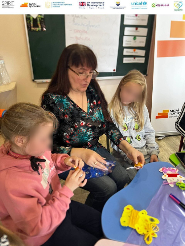 Дві дівчинки сидять з жінкою за столиком. Одна  дивиться їй на руки, інша - розгортає цукерку.