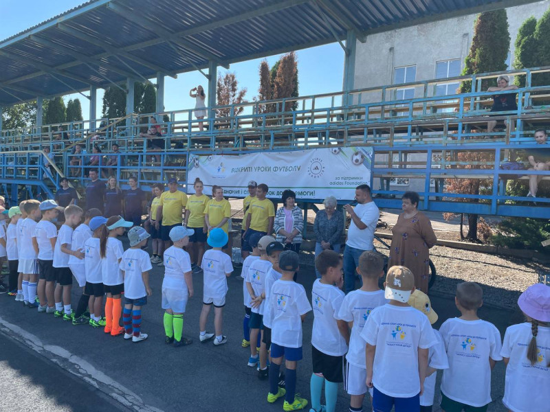 Діти в однакових  білих футболках з емблемою Всеукраїнської фундації «Захист прав дітей»  стоять перед трибуною і слухають виступ молодого чоловіка. Біля нього- групи з чоловіків і жінок у жовтих та синіх футболках.