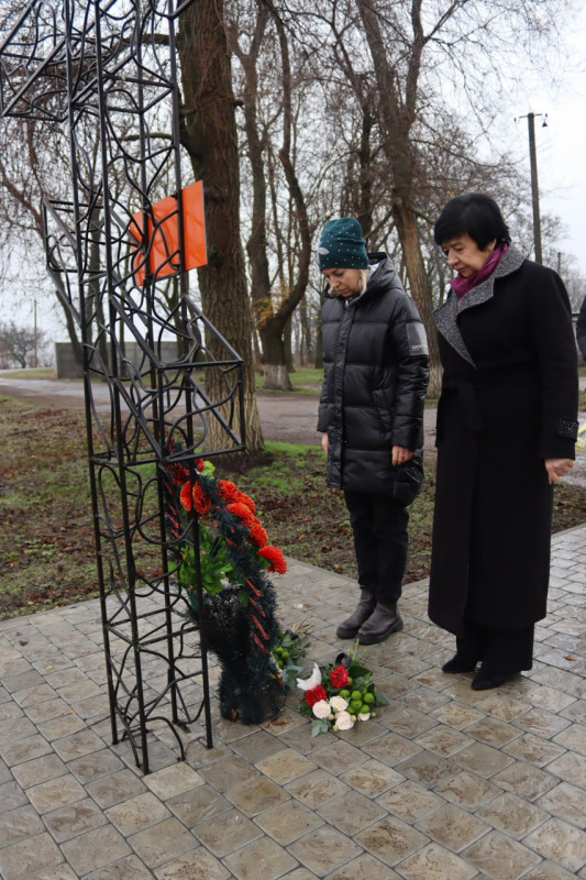 Начальник Синельниківської районної військової адміністрації Світлана Назарчук та секретар міської ради Олена Заіка стоять біля пам'ятного хреста, схиливши голови. Біля хреста  - квіти.