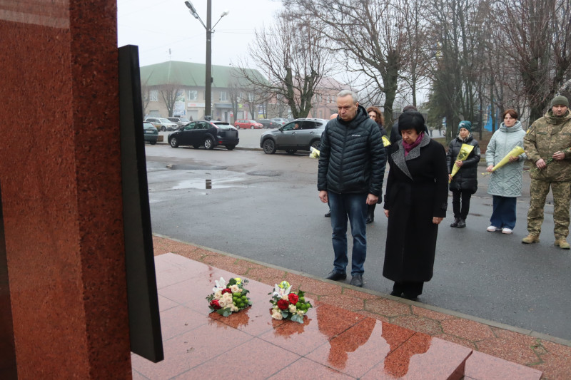 Міський голова Дмитро Зражевський, начальник Синельниківської районної військової адміністрації Світлана Назарчук стоять у хвилині мовчання перед пам'ятним знаком на честь полеглих Героїв сучасності. На підніжжі знаку - квіти.  За ними -  інші представники міської та районнної влади, військові з квітами. 