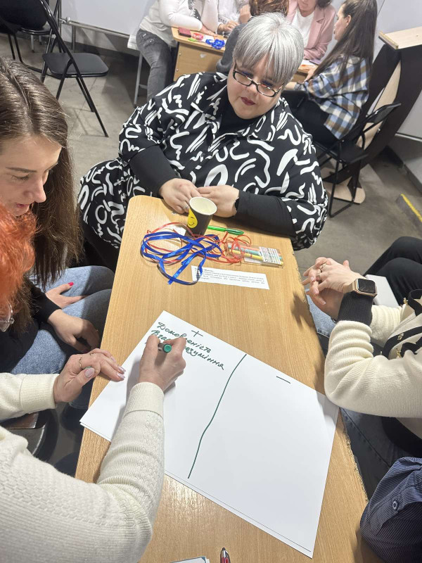 Група учасників заходу сидить за столом. Одна із учасниць пише на аркуші паперу текст. 